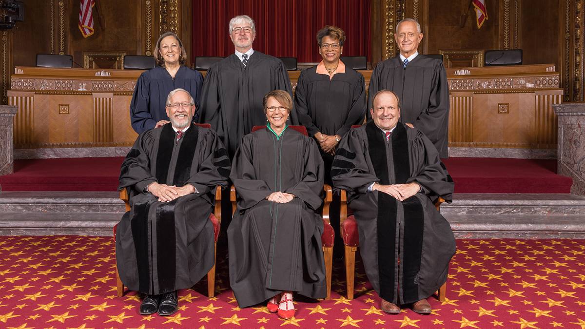 Image of seven men and women, all wearing black judicial robes, four standing and three seated, in the courtroom of the Thomas J. Moyer Ohio Judicial Center.